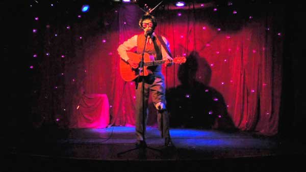 Musical comedian Elliot Mason live on stage at Madame Jojos dressed as an alien with an eyeball on his knee